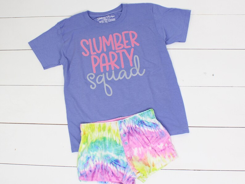 Może przedstawiać: Fioletowy T-shirt z napisem "slumber party squad" w r&oacute;żowych i szarych literach. T-shirt jest połączony z kr&oacute;tkimi spodenkami tie-dye w odcieniach r&oacute;żowego, niebieskiego, zielonego i ż&oacute;łtego.