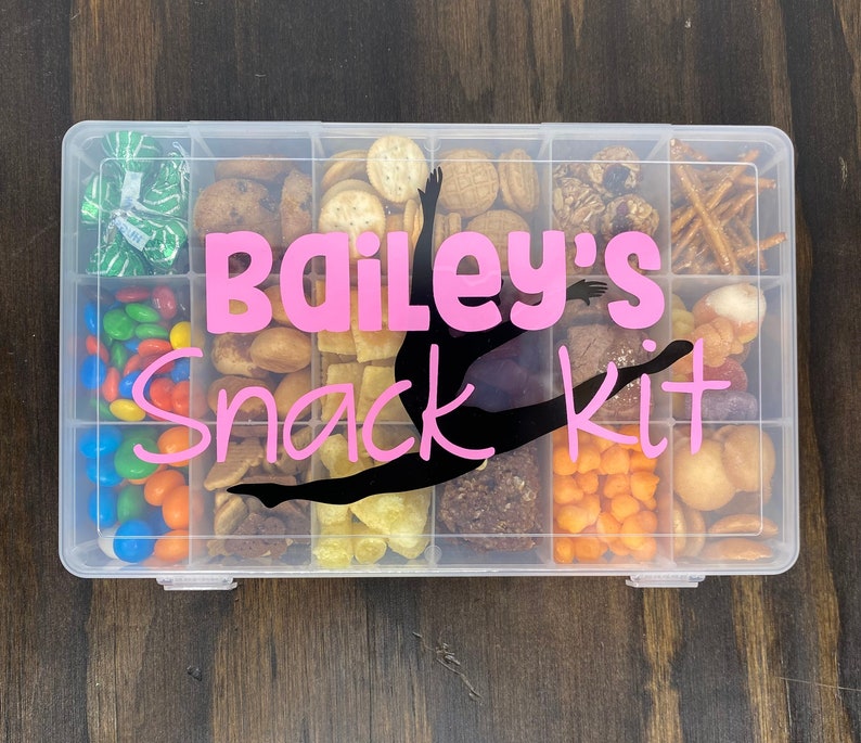 May include: A clear plastic container with multiple compartments filled with various snacks. The container has a pink label that says "Bailey's Snack Kit" with a black silhouette of a gymnast in the middle.