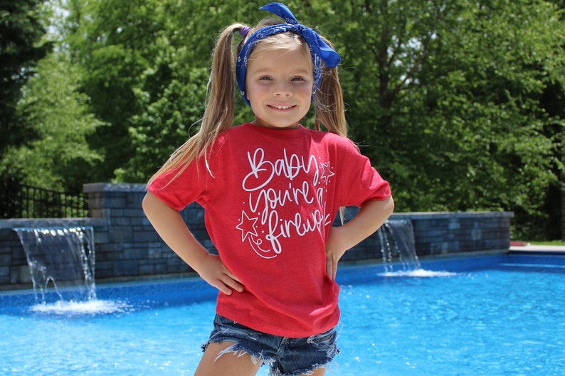 May include: A young girl wearing a red t-shirt with the text "Baby you're a firefly" in white. She is wearing a blue bandana in her hair and has her hands on her hips. She is standing in front of a swimming pool.