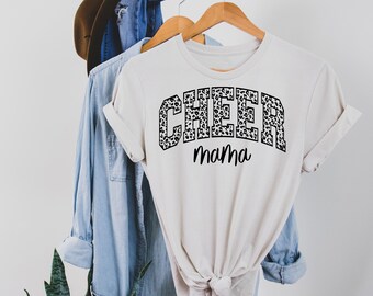 May include: A white t-shirt with a black graphic that says "CHEER" in a leopard print font and "Mama" underneath. The shirt is hanging on a wooden hanger.