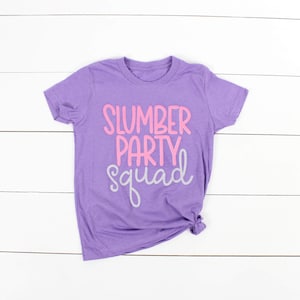 Może przedstawiać: Fioletowy T-shirt z napisem "Slumber Party Squad" w r&oacute;żowych i szarych literach.
