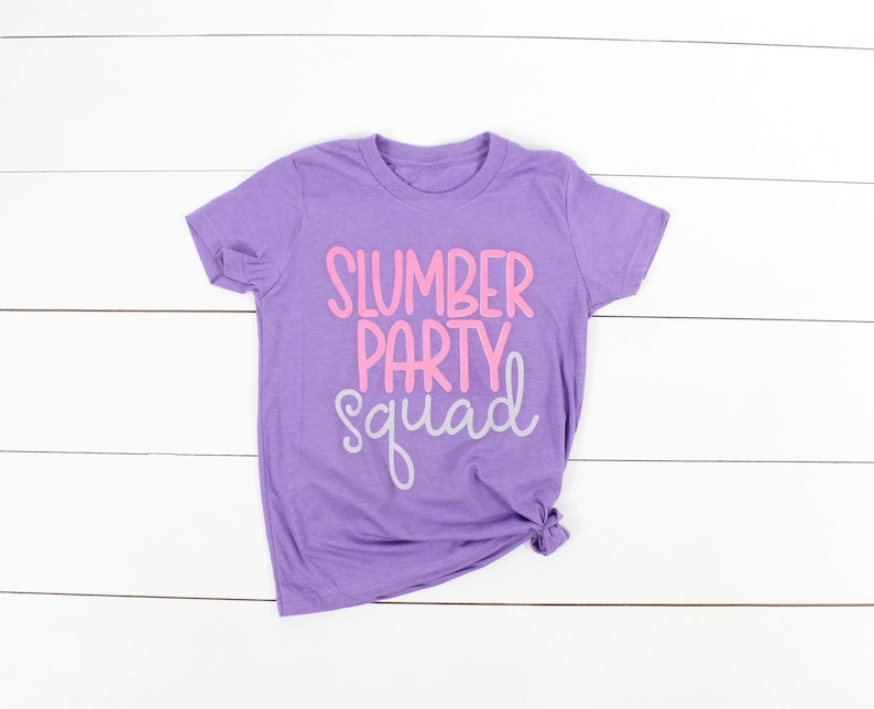 Może przedstawiać: Fioletowy T-shirt z napisem "Slumber Party Squad" w r&oacute;żowych i szarych literach.
