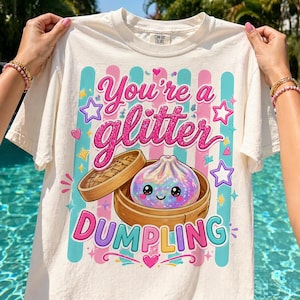 May include: Cream-colored t-shirt with a colorful graphic. The graphic features the text "You're a Glitter Dumpling" in pink and teal, along with a cartoon dumpling in a bamboo steamer, surrounded by stars and hearts.