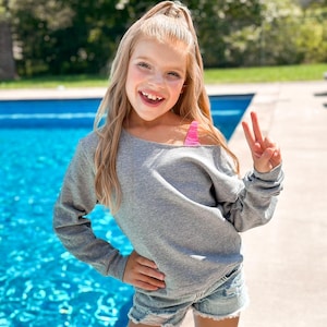 May include: A young girl wearing a gray off-the-shoulder sweatshirt, ripped denim shorts, and a pink swimsuit top. She is standing by a swimming pool and smiling at the camera.