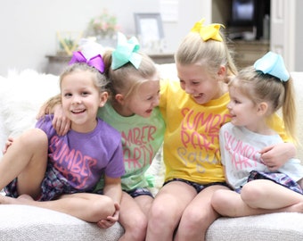 May include: Four girls wearing matching shirts with the text "Summer Party Squad" in pink and white lettering. The girls are sitting on a couch and smiling. They are wearing colorful hair bows.