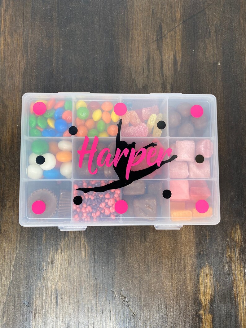 May include: A clear plastic container with multiple compartments filled with different types of candy. The container has a pink and black design with the name "Harper" and a silhouette of a gymnast.
