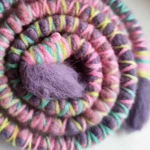 Op de afbeelding: Een close-up van een kleurrijke bol garen, met roze, paarse, groene en gele strepen. Het garen is in een spiraalvorm gewikkeld, met de uiteinden van het garen die twee kleine bollen vormen.