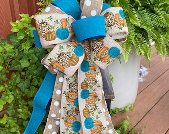May include: A decorative bow with a seasonal autumn theme. The bow features a burlap-style ribbon with white polka dots and a pumpkin print with leopard print accents. Teal blue ribbon provides contrast. The bow is attached to a black lantern with lit candles.
