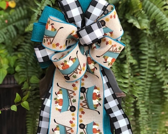 May include: A decorative autumn-themed bow made from various ribbons. The bow incorporates black and white buffalo check, teal, and beige ribbons. The beige ribbon displays a pattern of pumpkins in a wheelbarrow with autumn leaves. Suitable for seasonal decorations.