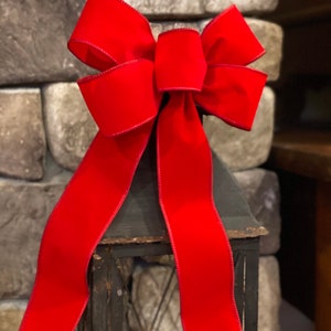 May include: A large, vibrant red bow with long, flowing tails. The bow is made of a velvety material and is placed atop a black metal lantern. The lantern has a distressed finish, and the background features a stone wall.