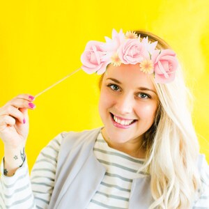 May include: A woman with blonde hair is wearing a light blue waistcoat over a white and light blue striped shirt. She is holding a flower crown prop with pink and white flowers. The background is yellow.