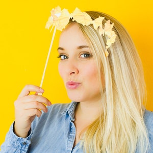 Op de afbeelding: Een vrouw met blond haar draagt een blauw denim shirt en houdt een stok vast met een papieren vlinderdecoratie erop. Ze kijkt naar de camera en heeft een lichte glimlach op haar gezicht. De achtergrond is felgeel.