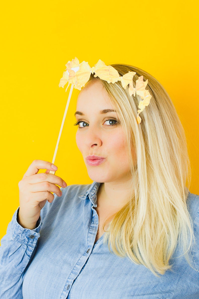 Op de afbeelding: Een vrouw met blond haar draagt een blauw denim shirt en houdt een stok vast met een papieren vlinderdecoratie erop. Ze kijkt naar de camera en heeft een lichte glimlach op haar gezicht. De achtergrond is felgeel.