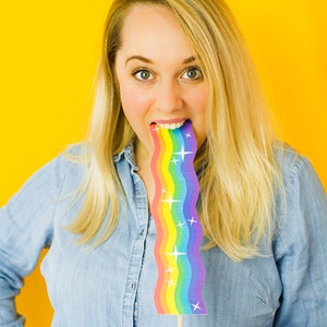 Op de afbeelding: Een vrouw met blond haar houdt een regenboogkleurig papieren rekwisiet voor haar mond. Het rekwisiet is golvend en heeft witte sterren erop. De achtergrond is geel.