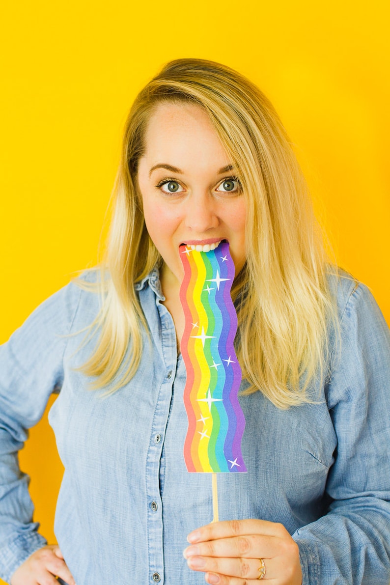Op de afbeelding: Een vrouw met blond haar houdt een regenboogkleurig papieren rekwisiet voor haar mond. Het rekwisiet is golvend en heeft witte sterren erop. De achtergrond is geel.