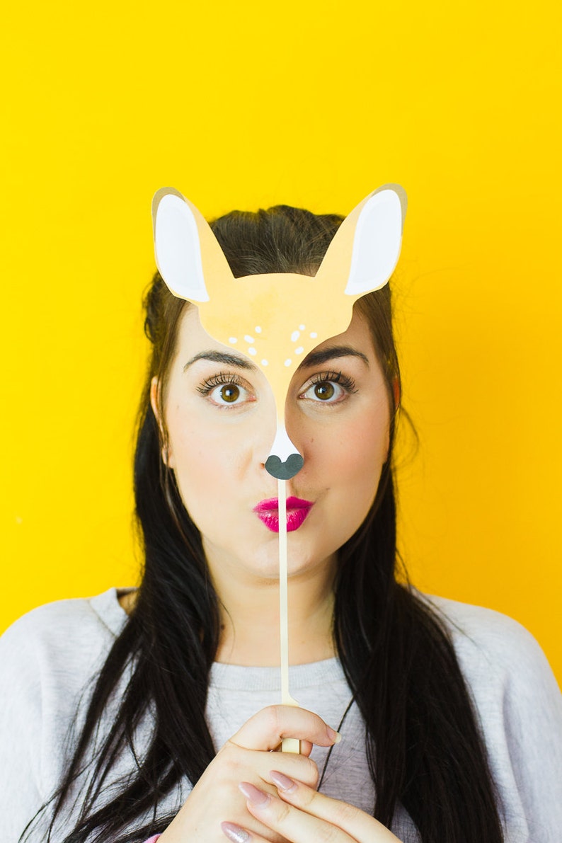 Op de afbeelding: Een vrouw houdt een papieren hertenmasker voor haar gezicht. Het masker is geel met bruine vlekken en zwarte ogen. De vrouw draagt een grijs shirt en heeft lang zwart haar.