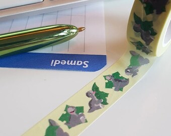Op de afbeelding: Een rol decoratieve washi-tape met een herhalend patroon van grijze dinosaurussen en groene bladeren op een lichtgele achtergrond. De tape is gedeeltelijk afgerold en toont het ontwerp. Een groene pen en een toetsenbord staan op de achtergrond.