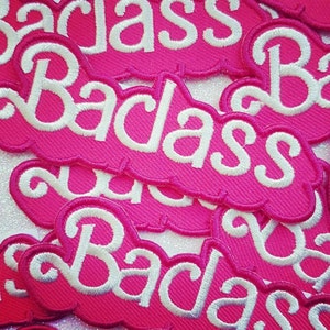 Op de afbeelding: Een verzameling roze geborduurde patches met het woord "Badass" in witte letters. De patches hebben een wolkenvorm en zijn dicht op elkaar geplaatst, waardoor het herhaalde ontwerp wordt benadrukt.