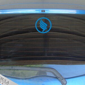 K&ouml;nnte beinhalten: Ein blaues Auto mit einem blauen und wei&szlig;en Logoaufkleber auf der Heckscheibe. Das Logo ist ein stilisierter Vogel mit ausgebreiteten Fl&uuml;geln. Der Text "RED NEBULA STUDIOS" ist in der linken unteren Ecke des Bildes sichtbar.
