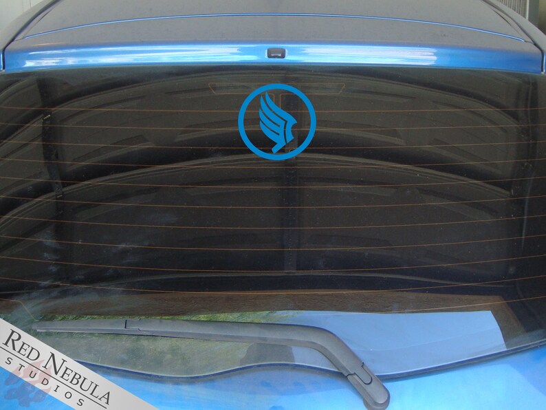 K&ouml;nnte beinhalten: Ein blaues Auto mit einem blauen und wei&szlig;en Logoaufkleber auf der Heckscheibe. Das Logo ist ein stilisierter Vogel mit ausgebreiteten Fl&uuml;geln. Der Text "RED NEBULA STUDIOS" ist in der linken unteren Ecke des Bildes sichtbar.