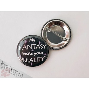 May include: A black button with a white design that reads "My Fantasy beats your Reality" with pink stars. The button has a silver pin back.