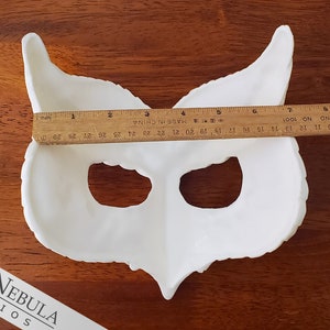 May include: White owl mask made of plaster or resin. The mask is approximately 33 cm wide and has two eye holes. The mask is on a wooden surface.