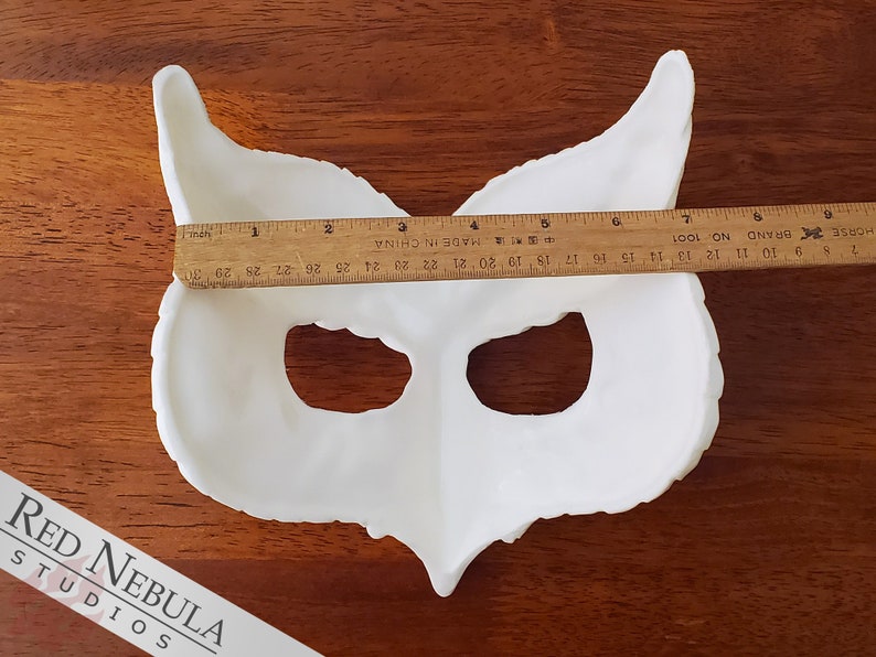 May include: White owl mask made of plaster or resin. The mask is approximately 33 cm wide and has two eye holes. The mask is on a wooden surface.