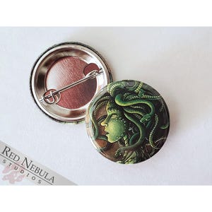 May include: A round button with a green and brown image of a woman with snakes for hair. The woman's face is visible and she has a green and brown snake wrapped around her head. The button is a 1 inch button.