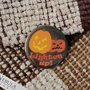 May include: A black button with a Halloween theme. Two orange pumpkins with jack-o'-lantern faces are on the button. The text "Lighten Up!" is written in white on the button.