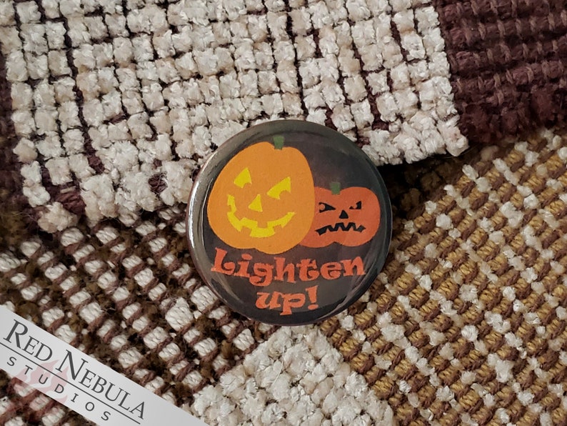 May include: A black button with a Halloween theme. Two orange pumpkins with jack-o'-lantern faces are on the button. The text "Lighten Up!" is written in white on the button.