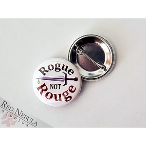 May include: A white button with a purple dagger and the text "Rogue Not Rouge" in red and black lettering.