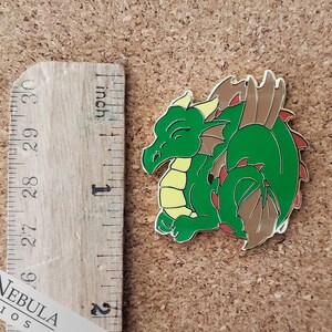 May include: A green dragon enamel pin with yellow and brown accents. The dragon is lying down with its eyes closed. The pin is approximately 1 inch in length. The text "Red Nebula Studios" is visible.