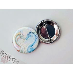May include: A button featuring a watercolor illustration of two dragons forming a heart shape. The dragons are blue and orange with a pink and white background.