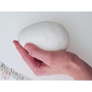 May include: A white, smooth, egg-shaped object is held in a person's hand. The object is about the size of a large chicken egg.