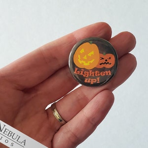 May include: A round button with a black background featuring two orange pumpkins with jack-o'-lantern faces. The larger pumpkin has a happy expression, while the smaller pumpkin has a grumpy expression. The text "Lighten up!" is written in white letters below the pumpkins.