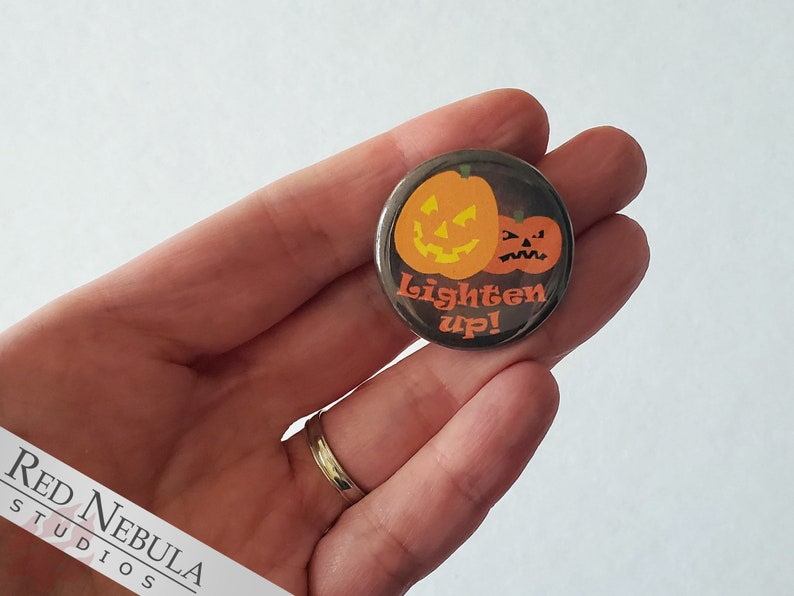May include: A round button with a black background featuring two orange pumpkins with jack-o'-lantern faces. The larger pumpkin has a happy expression, while the smaller pumpkin has a grumpy expression. The text "Lighten up!" is written in white letters below the pumpkins.