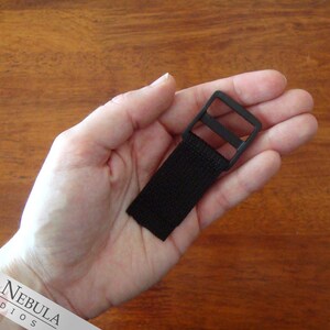 Op de afbeelding: Zwarte plastic gesp met een zwarte nylon band eraan bevestigd. De gesp is rechthoekig met een enkele gleuf voor de band.