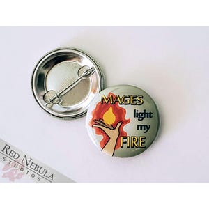 May include: A round button with a silver back and a colorful front. The front features a hand holding a flame with the text "MAGES light my FIRE".