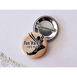 May include: A beige button with a black and white illustration of a foot wearing a shoe with the text "Fen'Harel ver na" on it.