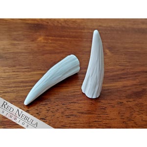 May include: Two white, textured, plastic horns. The horns are curved and pointed. They are on a wooden surface.