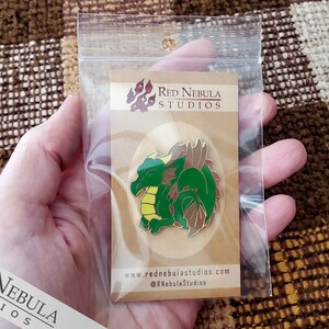 May include: A green and brown dragon enamel pin with yellow accents, displayed on a cardboard backing inside a clear plastic bag. The pin features the text "Red Nebula Studios" and website information.