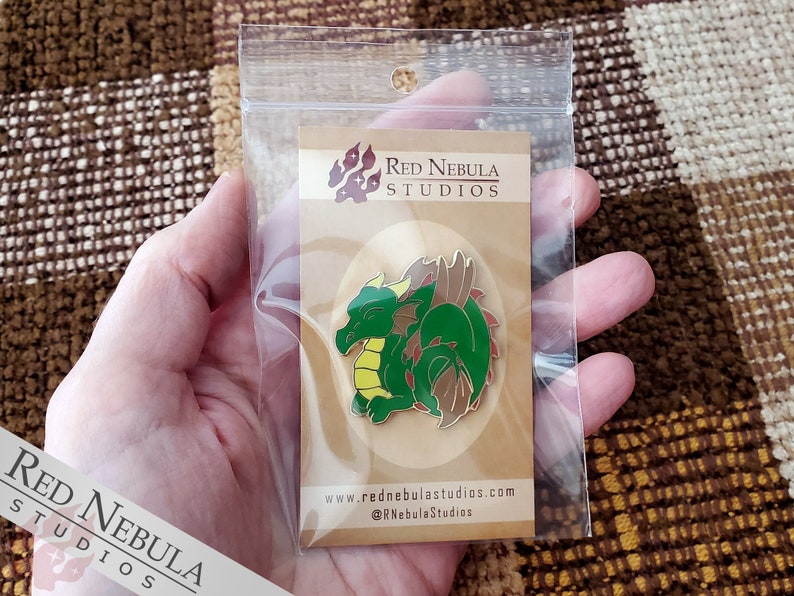 May include: A green and brown dragon enamel pin with yellow accents, displayed on a cardboard backing inside a clear plastic bag. The pin features the text "Red Nebula Studios" and website information.