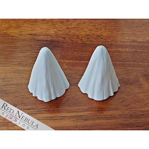 May include: Two white ghost figurines on a wooden surface. The ghosts are made of a smooth, white material and have a flowing, ethereal appearance.
