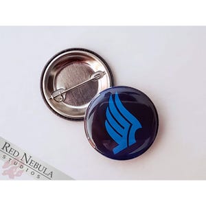 May include: A blue and black button with a stylized wing design. The button is 1 inch in diameter and has a metal back.
