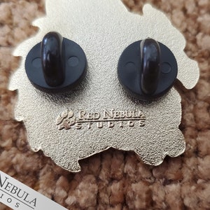 May include: Close-up of the back of a gold-colored enamel pin with two black pin backs. The pin has a unique shape and features the text "Red Nebula Studios". The pin is on a brown, textured surface.