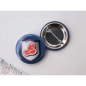 May include: A blue button with a white shield and a red steak inside. The back of the button is silver with a pin.