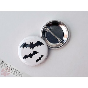 May include: A white button with a black silhouette of three bats. The button has a metal back with a safety pin.