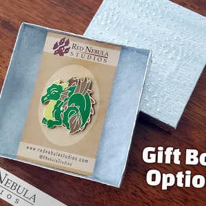 May include: A green and brown dragon enamel pin displayed in a silver gift box. The pin features a smiling dragon with yellow accents. The gift box is open, revealing the pin on a brown card with the text "Red Nebula Studios".