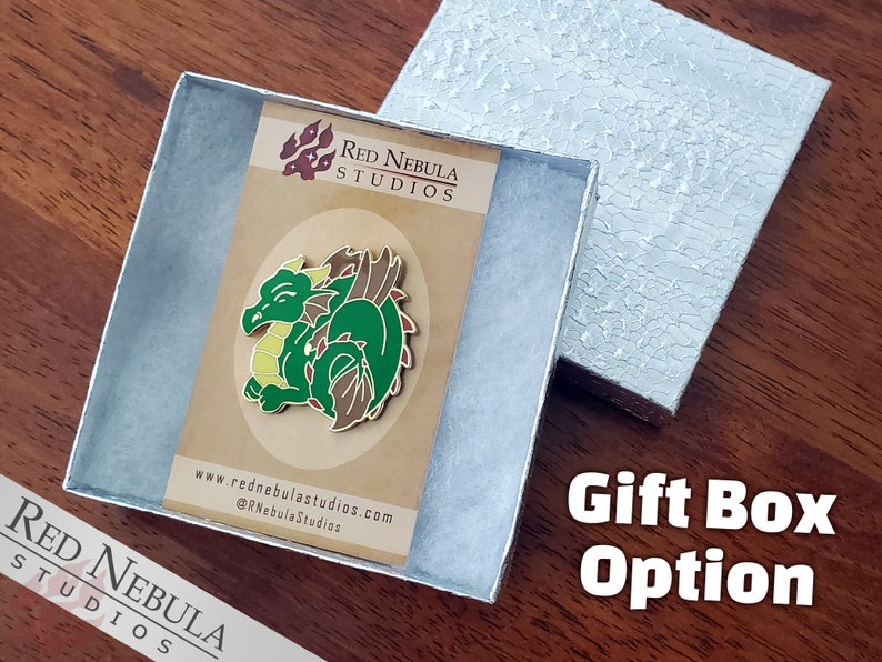 May include: A green and brown dragon enamel pin displayed in a silver gift box. The pin features a smiling dragon with yellow accents. The gift box is open, revealing the pin on a brown card with the text "Red Nebula Studios".