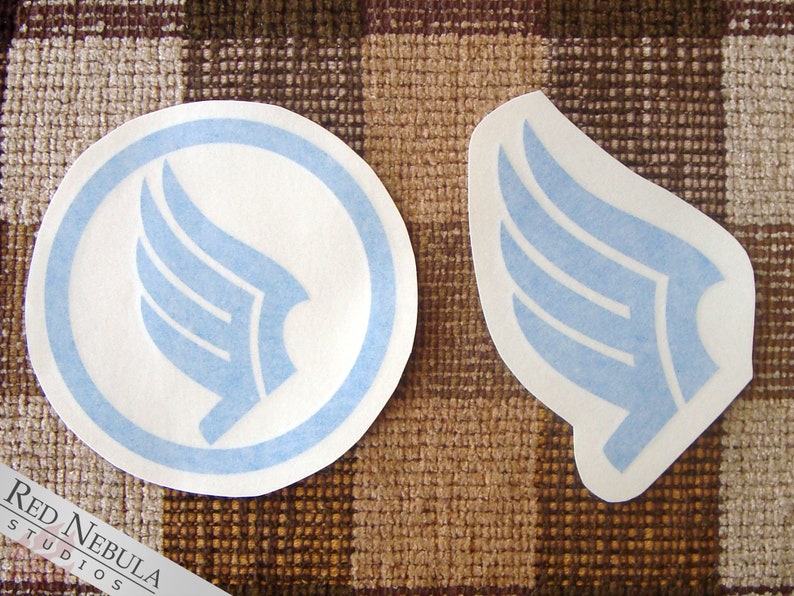 K&ouml;nnte beinhalten: Zwei blaue und wei&szlig;e Aufkleber mit einem stilisierten Fl&uuml;geldesign. Ein Aufkleber ist ein Kreis mit einem blauen Rand und der andere ist eine Fl&uuml;gelform.